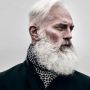 An older man with a full, white beard and short white hair looks to the side. He is wearing a dark coat and a patterned scarf, set against a neutral gray background.