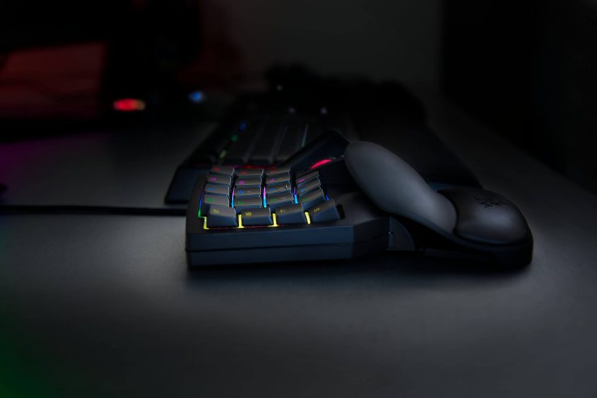 A close-up of a one-handed gaming keypad with RGB backlit keys and an ergonomic wrist rest, placed on a dark desk with a blurred keyboard in the background.