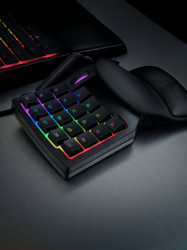 A compact gaming keypad with RGB backlit keys sits on a desk next to a black computer mouse and a keyboard with rainbow-colored lighting in the background.