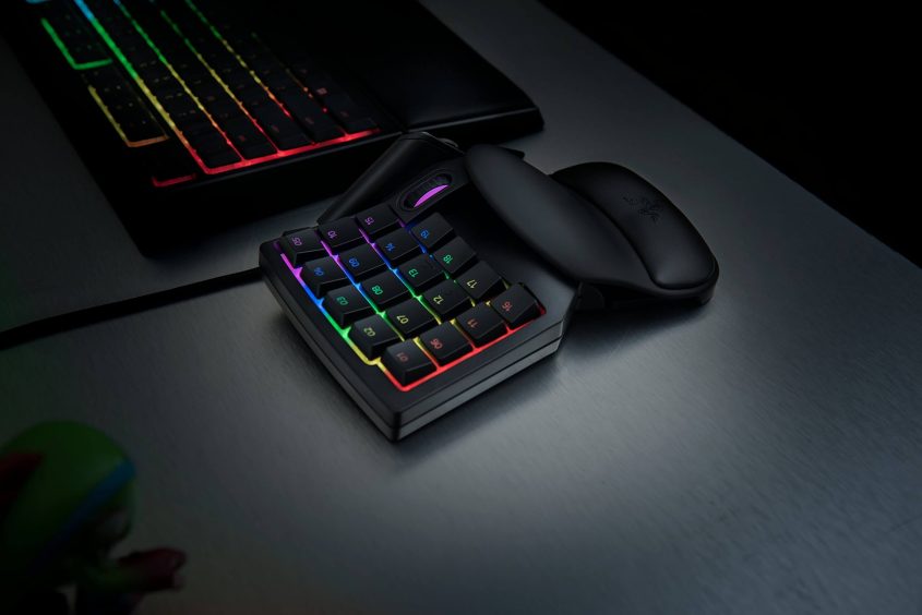 A compact gaming keypad with RGB backlit keys sits on a desk next to a black computer mouse and a keyboard with rainbow-colored lighting in the background.