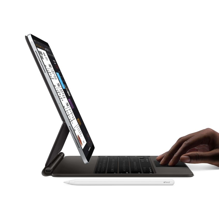 A person uses an iPad attached to a keyboard case, with an Apple Pencil resting on the surface in front of them. The iPad screen displays multiple open apps.