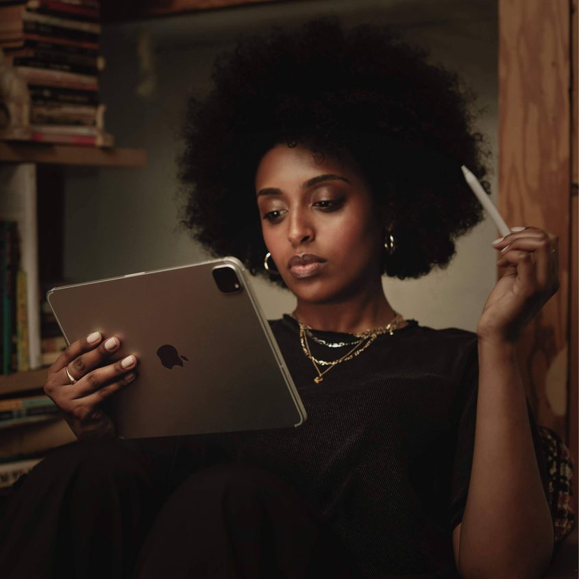 A woman with curly hair sits by bookshelves, holding a tablet in one hand and a stylus in the other, looking thoughtfully at the screen. She wears gold jewelry and a black shirt.
