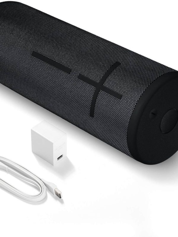 A cylindrical black portable Bluetooth speaker with large plus and minus volume buttons, shown next to a white USB charging cable and a white USB wall adapter.