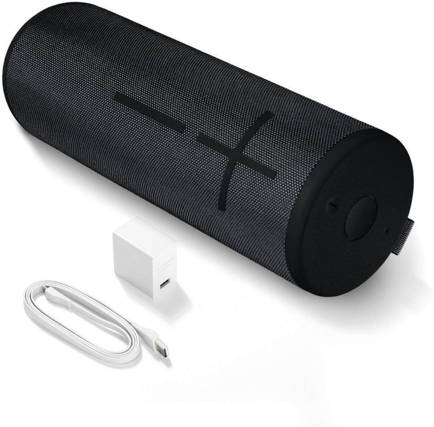 A cylindrical black portable Bluetooth speaker with large plus and minus volume buttons, shown next to a white USB charging cable and a white USB wall adapter.