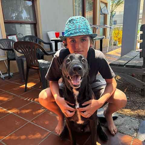 A boy wearing a blue patterned hat crouches on a patio, hugging and smiling with a happy black dog. Outdoor chairs, tables, and plants are visible in the background, adding to the cheerful Christmas atmosphere.