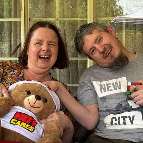 A woman and a man sit smiling together outside in front of glass doors and curtains. The woman holds a teddy bear wearing a Hot Cares shirt, adding a touch of Christmas cheer, while the man proudly holds a Rubiks Cube.