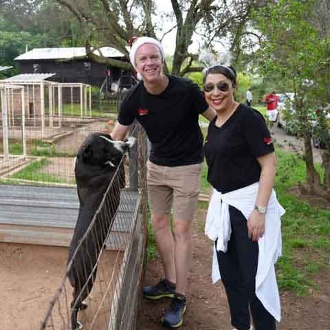 Two people in black shirts stand by a fenced enclosure during the Toy Run, smiling at the camera. One pets a black and white dog standing on its hind legs against the fence. Trees and more enclosures are in the background.
