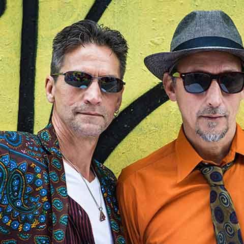 Two men wearing sunglasses pose in front of a yellow wall with black graffiti, channeling OMD style. One sports a colorful patterned jacket, white shirt, and necklace; the other wears a gray hat, orange shirt, and patterned tie.