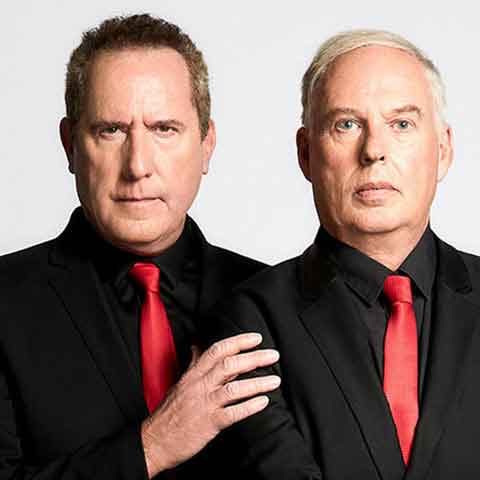 Two men from OMD wear black suits, black shirts, and red ties as they stand side by side against a plain light background. One rests his hand on the others shoulder, both displaying serious expressions.