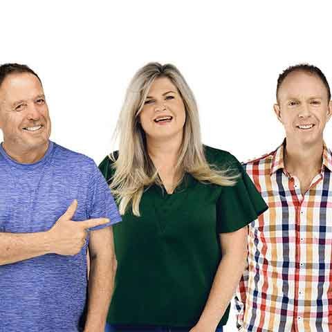 Three adults stand together smiling against a white background, perhaps after a successful appointment. The person on the left wears a blue shirt, the middle has long blonde hair and a green blouse, while the person on the right sports a colorful plaid shirt.