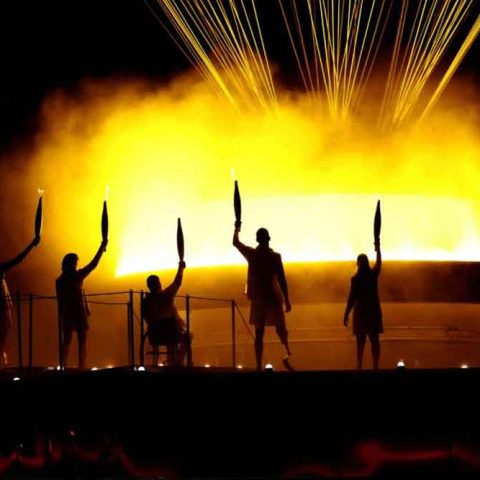 Five silhouetted figures hold torches aloft in front of a brightly lit cauldron, yellow flames and beams of light illuminating a ceremony that unfolds dramatically against the dark background.