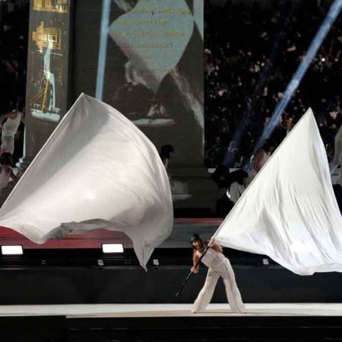 Two performers dressed in white wave large white flags on a brightly lit stage during a ceremony, with a crowd in the background and spotlights shining across the scene.