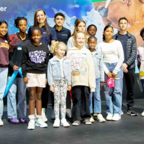 A group of 15 children and teenagers stand together on a stage, smiling at the camera. Dressed casually against a colorful background, they capture a moment to remember at what appears to be an event or presentation.
