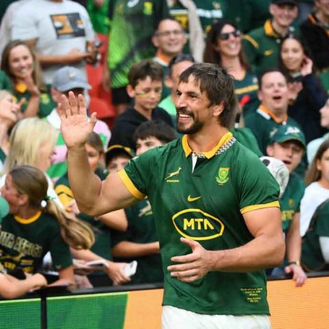 A rugby player in a green Springbok jersey waves and smiles at cheering fans in a crowded stadium, with many spectators wearing matching team colors and showing excitement.