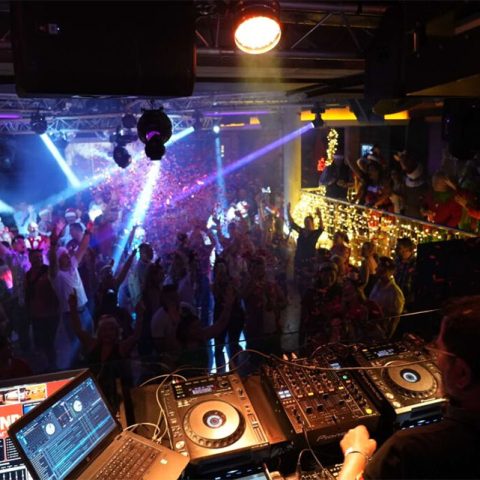 A DJ performs on stage with mixing equipment, spinning Old Skool beats while a large crowd dances under colorful lights in a lively nightclub setting. Bright spotlights and decorations add to the festive atmosphere.