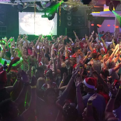 A large crowd of people at a lively indoor Old Skool party or concert, many wearing Santa hats and festive outfits, with hands raised and colorful lights illuminating the scene.