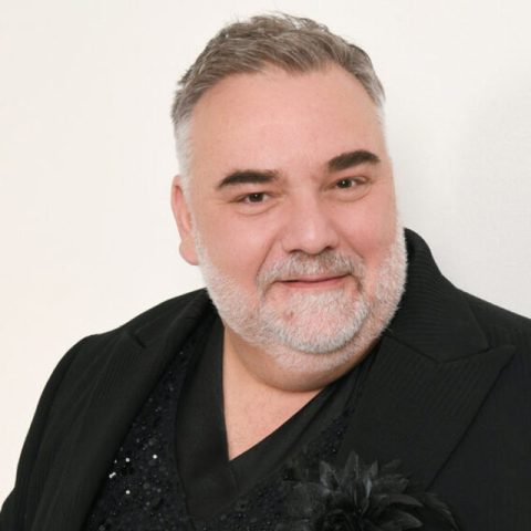 A middle-aged man with short gray hair and a beard smiles at the camera. He is wearing a black blazer with a Christmas-themed floral decoration on the lapel. The background is plain white.