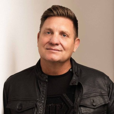 A man with short, styled brown hair is wearing a black leather jacket and a black shirt, standing indoors against a white wall and smiling gently at the camera, capturing a subtle hint of Christmas warmth.