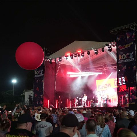 A large crowd gathers at Marks Park to watch a live outdoor concert at night. The stage glows with red lights, a band performs, and a big red balloon floats above the audience. Event banners proudly display “ROCK.”.