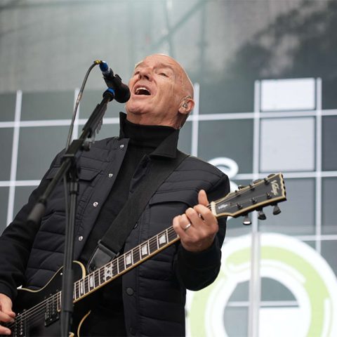 A bald man wearing a black jacket plays an electric guitar and sings into a microphone on stage at Marks Park, with a geometric patterned background behind him.