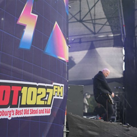 A large stage banner for HOT 102.7 FM is in the foreground at Marks Park, with the slogan Playing Joburg’s Best Old Skool and R&B. In the background, a musician plays guitar on stage under vibrant lights and scaffolding.