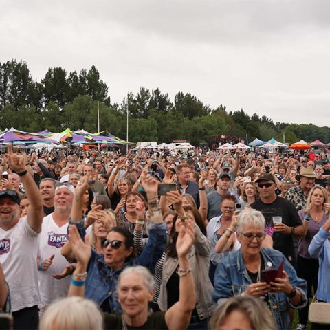 A large crowd at Marks Park raises their hands and smiles at an outdoor event. Colorful tents and trees fill the background under a cloudy sky, as many people enjoy a lively concert or festival.