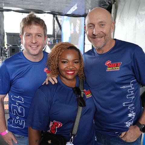 Three people wearing matching blue Hot 102.7 FM t-shirts smile and pose together backstage at Marks Park, with equipment and a stage visible in the background.
