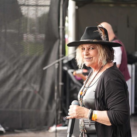 A woman wearing a black hat with a feather, layered necklaces, and a black cardigan stands on an outdoor stage at Marks Park, holding a microphone and smiling at the camera. Musicians and instruments are visible in the background.
