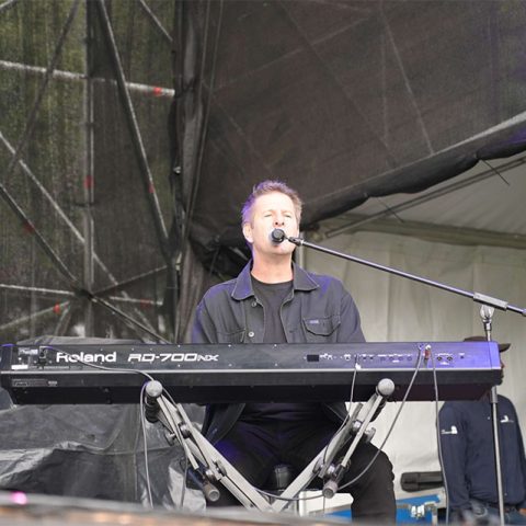 A man wearing a black jacket plays a Roland electric keyboard and sings into a microphone on an outdoor stage at Marks Park, with stage equipment and a white tent visible in the background.