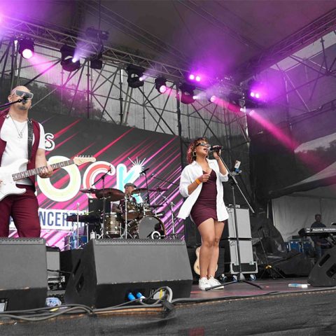 A band performs on an outdoor stage at Marks Park, illuminated by colorful lights and a vibrant backdrop. Three members stand at the front singing and playing instruments, while a drummer and keyboard player are visible in the background.