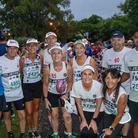 A group of runners in race bibs and athletic gear poses together at Marks Park before a race, smiling at the camera. Trees and other participants are visible in the background.