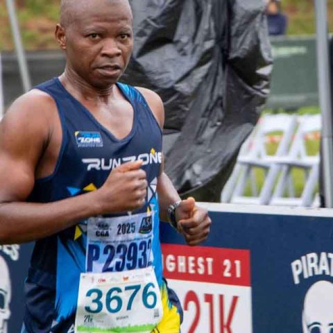 A focused runner wearing a blue Run Zone vest and race bib number 3676 nears the finish line at Marks Park, with event signage and other participants visible in the background.