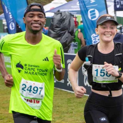 Two runners, a man in a bright green shirt and a woman in black athletic gear, smile as they run side by side during a race at the Cape Town Marathon near Marks Park, with banners and spectators cheering in the background.