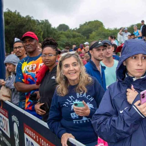 A group of people stand outdoors near a barrier with a pirate skull logo at Marks Park, some smiling, others holding phones, dressed in casual and rain gear, with a crowd and trees in the background.