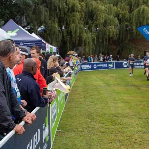 Crowd of spectators stand behind a barrier at Marks Park, watching and cheering as several runners approach the finish line of an outdoor race on a grassy course with banners and trees in the background.