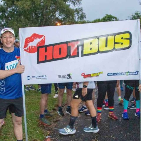 Two men in athletic gear hold a banner reading HOT BUS with a red lipstick mark, standing among a group of runners at an outdoor race event in Marks Park. Trees and grass are visible in the background.