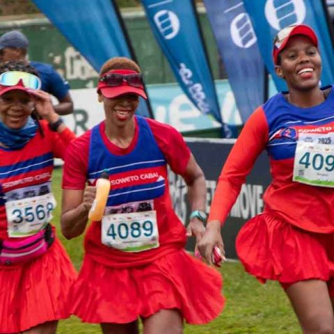 Three women wearing matching red and blue athletic outfits and skirts run together in a race at Marks Park, smiling and holding hands. They have race bibs numbered 3568, 4089, and 4096, with banners and other runners visible in the background.