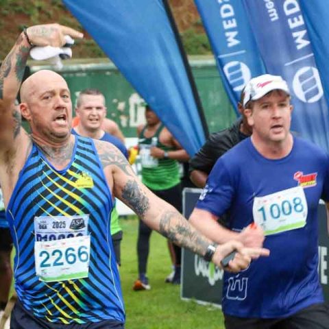 A group of runners approach the finish line at Marks Park during a race, with one bald man in a striped blue tank top raising his arm and another man in a blue shirt behind him, both wearing race bibs and looking determined.