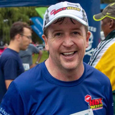 A smiling man wearing a blue race shirt and a white cap stands outdoors at Marks Park after a race, with other participants and colorful banners visible in the background.