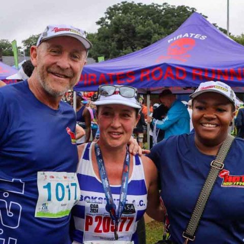 Three smiling people stand together at an outdoor running event at Marks Park, wearing bibs and athletic gear. Tents with “PIRATES ROAD RUNNING” are visible in the background on a grassy field.