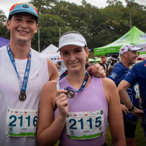 Two smiling runners wearing race bibs and medals pose together at the Marks Park outdoor event, with tents and other participants in the background. The woman proudly holds up her medal.