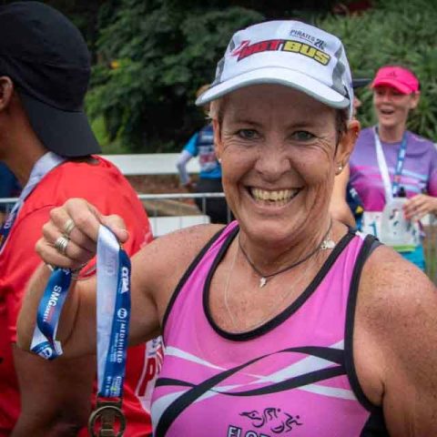 A smiling woman in a pink athletic outfit holds up a finisher’s medal at an outdoor race event in Marks Park. Other participants wearing race gear and medals are visible in the background.