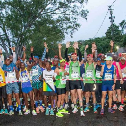 A large group of marathon runners stand at the starting line in Marks Park, smiling and raising their arms in excitement. They wear colorful athletic gear and numbered race bibs, with trees and a cloudy sky in the background.