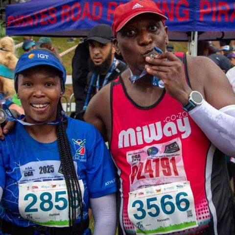 Two marathon runners smile after finishing at Marks Park, proudly holding their medals. The woman on the left in blue and the man on the right in red display race numbers 285 and 3566, with other runners in the background.