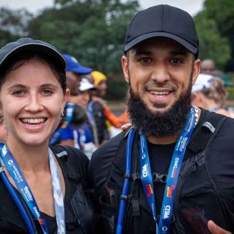 Two smiling runners wearing medals and black sports caps pose for a photo after a race at Marks Park, with other participants and greenery in the blurred background.