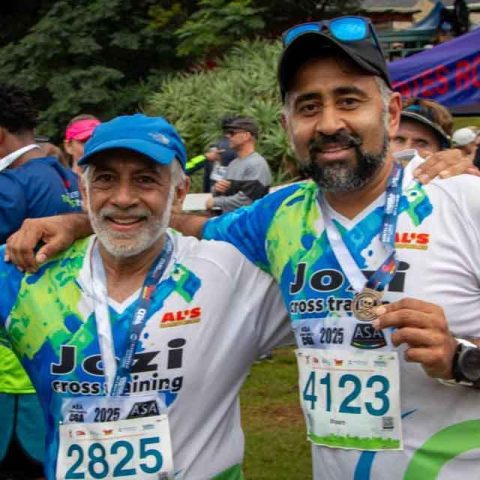 Two men wearing running medals and race bibs pose and smile after a race at Marks Park, with arms around each other. Other runners and a blue event tent are visible in the background.