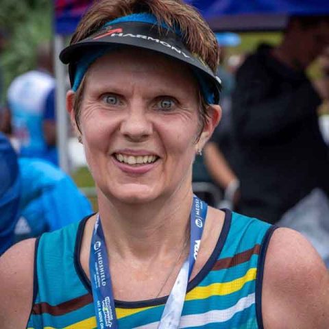 A smiling woman wearing a blue striped athletic tank top, a medal, and a visor stands outdoors among other people at what appears to be a race or sporting event at Marks Park.