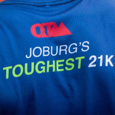 Back of a blue athletic shirt with the OTM logo and text reading JOBURGS TOUGHEST 21K in white, green, and blue lettering, celebrating the challenge at Marks Park.