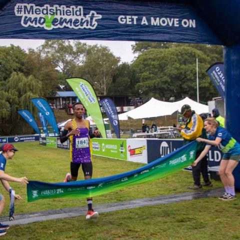 A runner in a purple outfit crosses the finish line at Marks Park, breaking the tape as two event staff hold the ends. The finish arch displays #Medshield Movement and Pirates, with outdoor event banners visible.