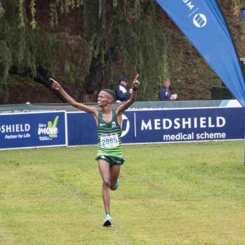 A runner in a green uniform crosses the finish line with arms raised in victory at Marks Park, while a photographer captures the moment. Event sponsor banners are visible in the background.